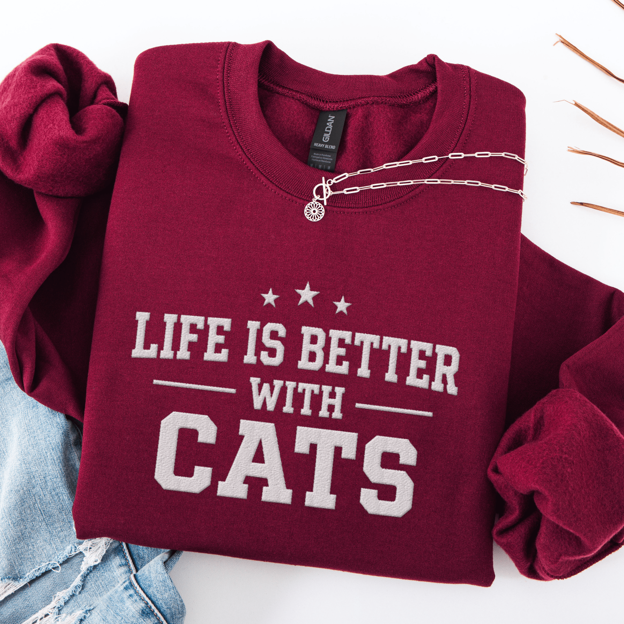 Maroon cat embroidered sweatshirt with “Life Is Better With Cats” text, folded flat on light denim