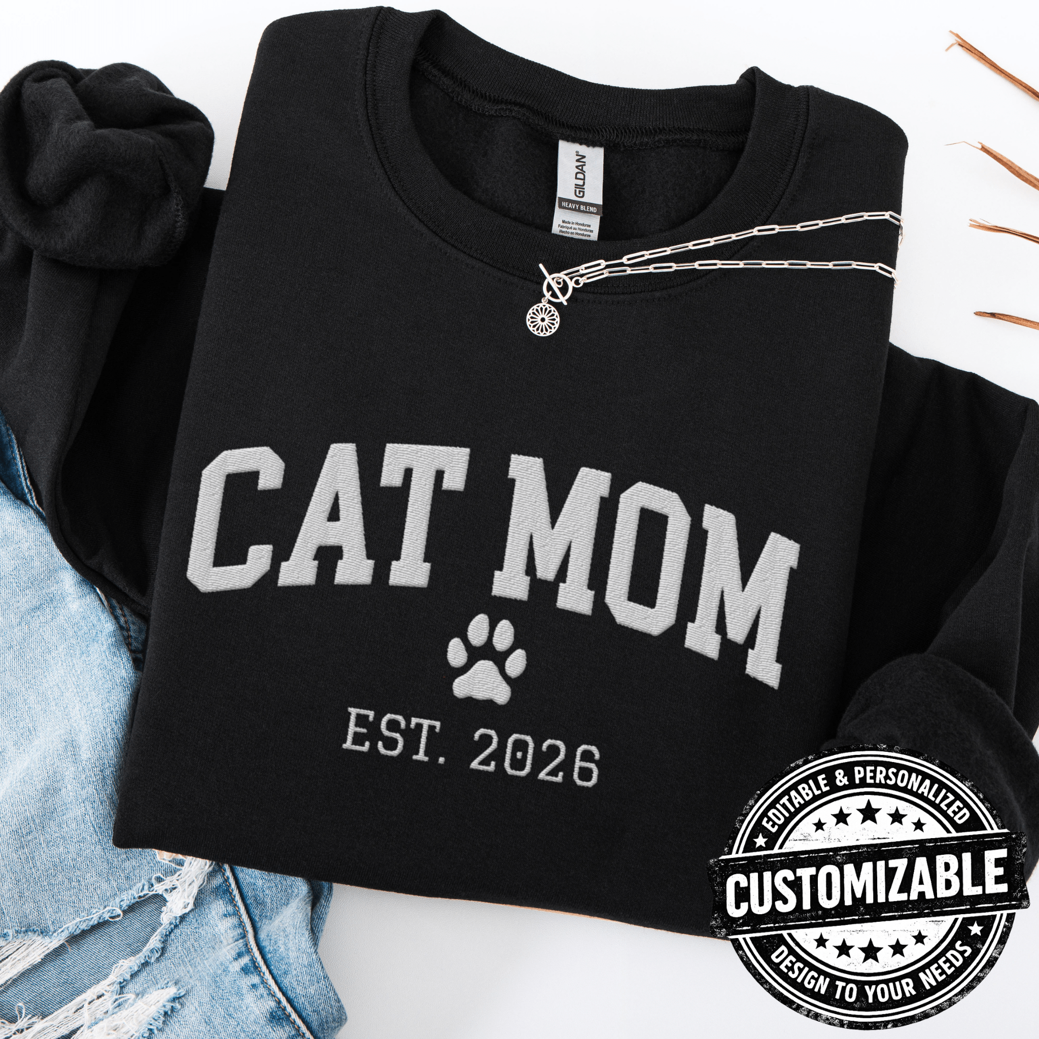 Black customizable cat mom embroidered sweatshirt with paw print and custom year detail, folded flat on light denim