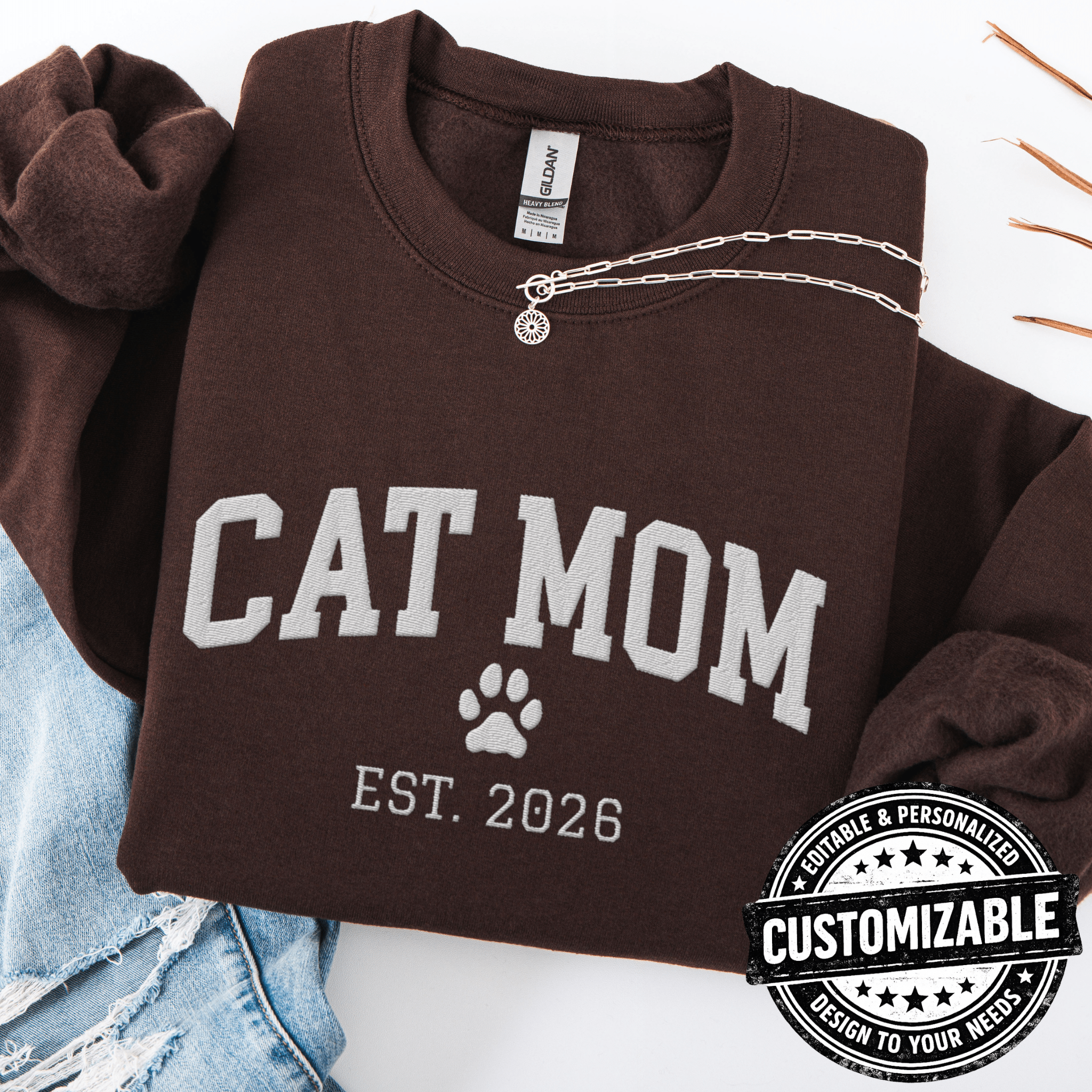 Dark Chocolate customizable cat mom embroidered sweatshirt with paw print and custom year detail, folded flat on light denim