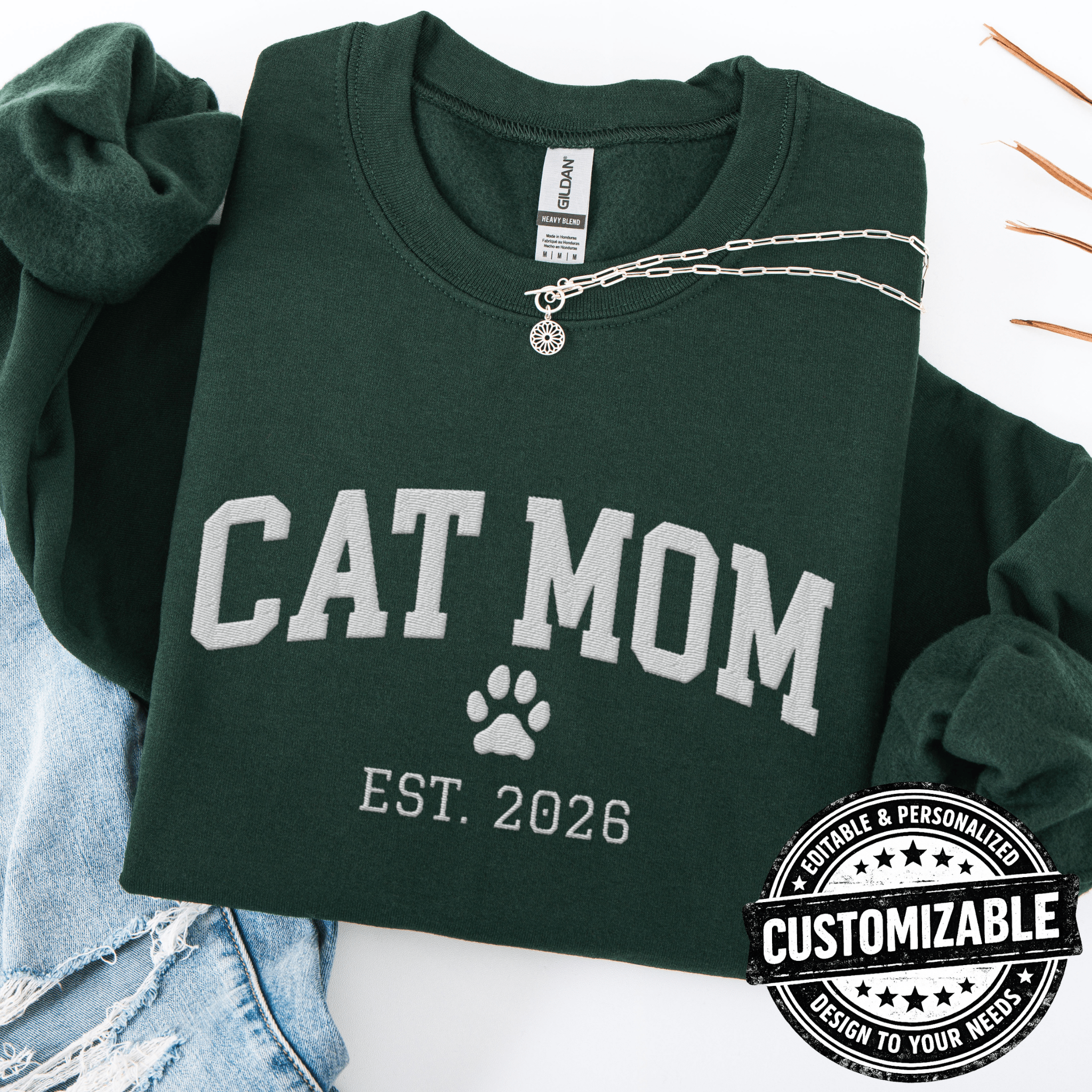 Forest Green customizable cat mom embroidered sweatshirt with paw print and custom year detail, folded flat on light denim