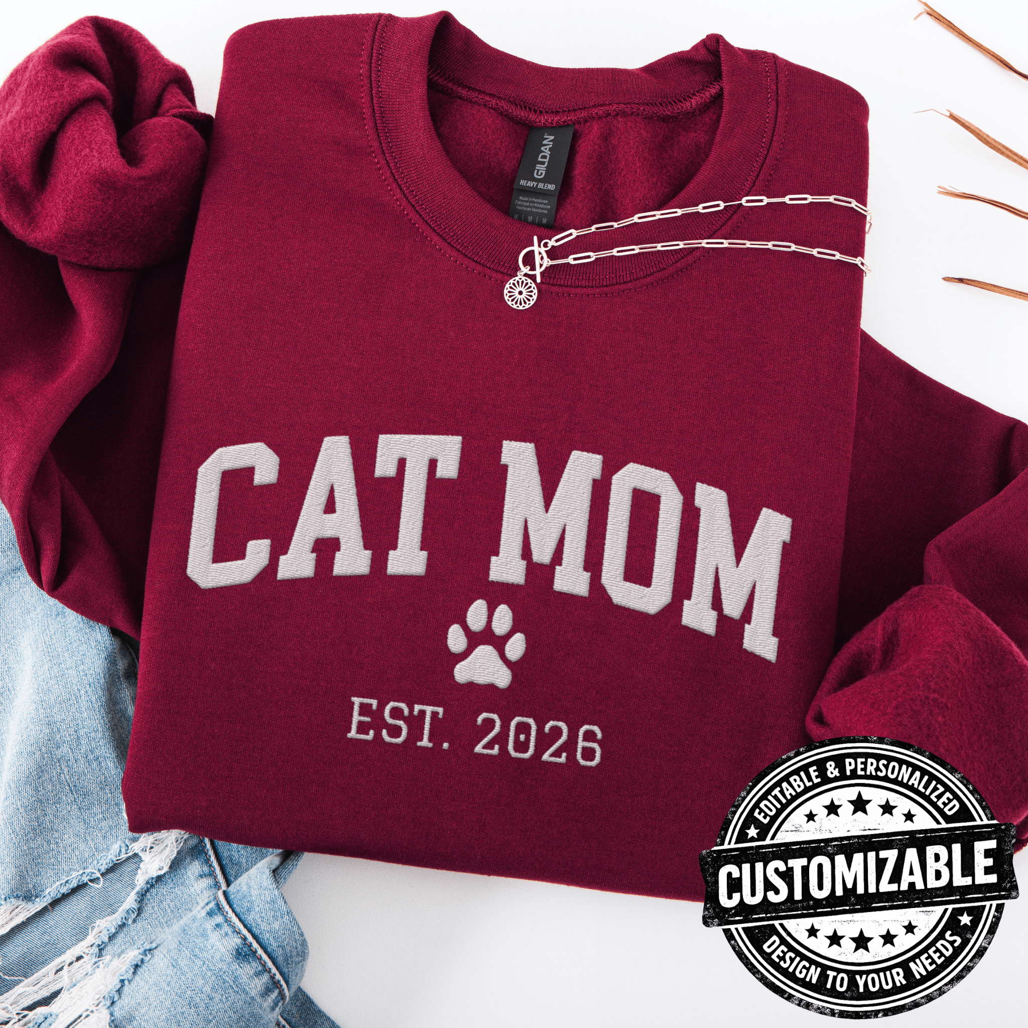 Maroon customizable cat mom embroidered sweatshirt with paw print and “Est. 2025” detail, folded flat on light denim