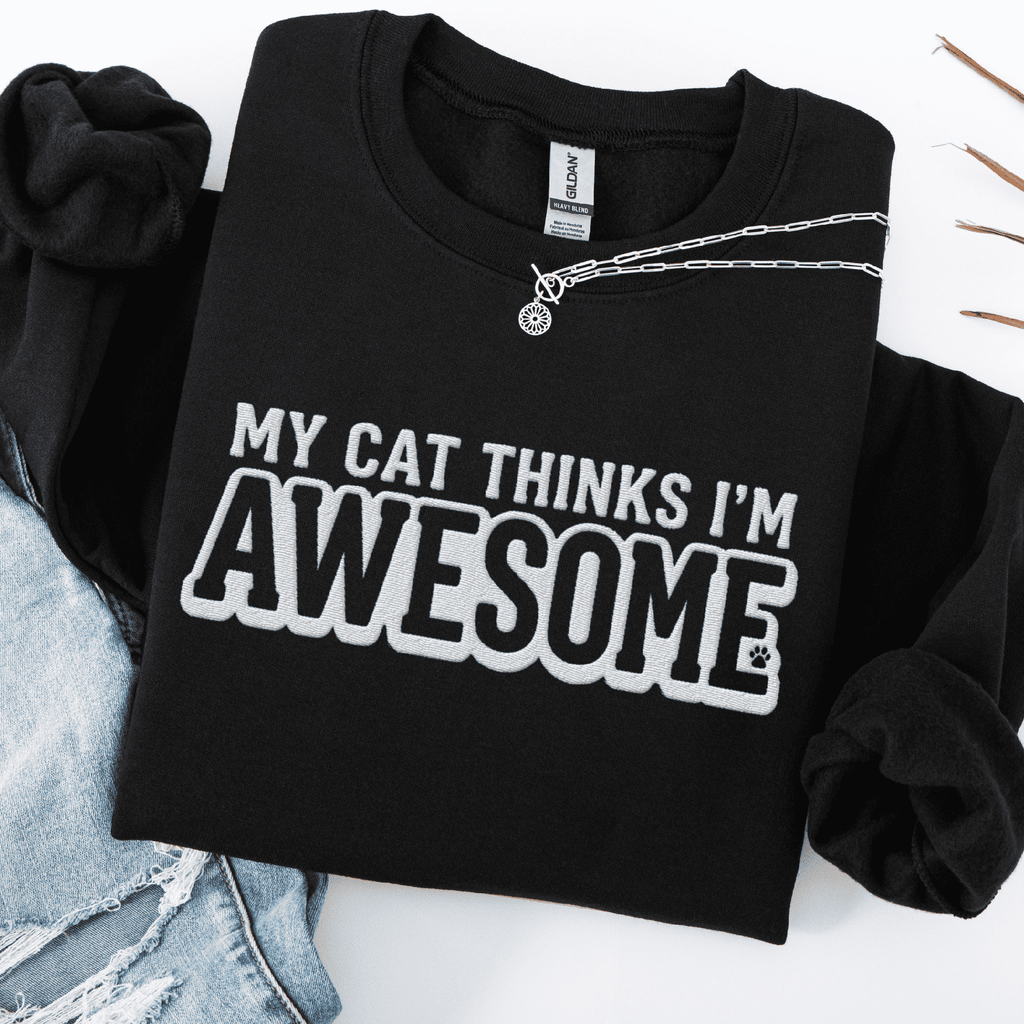 Black cat embroidered sweatshirt with “My Cat Thinks I’m Awesome” text, folded flat on light denim