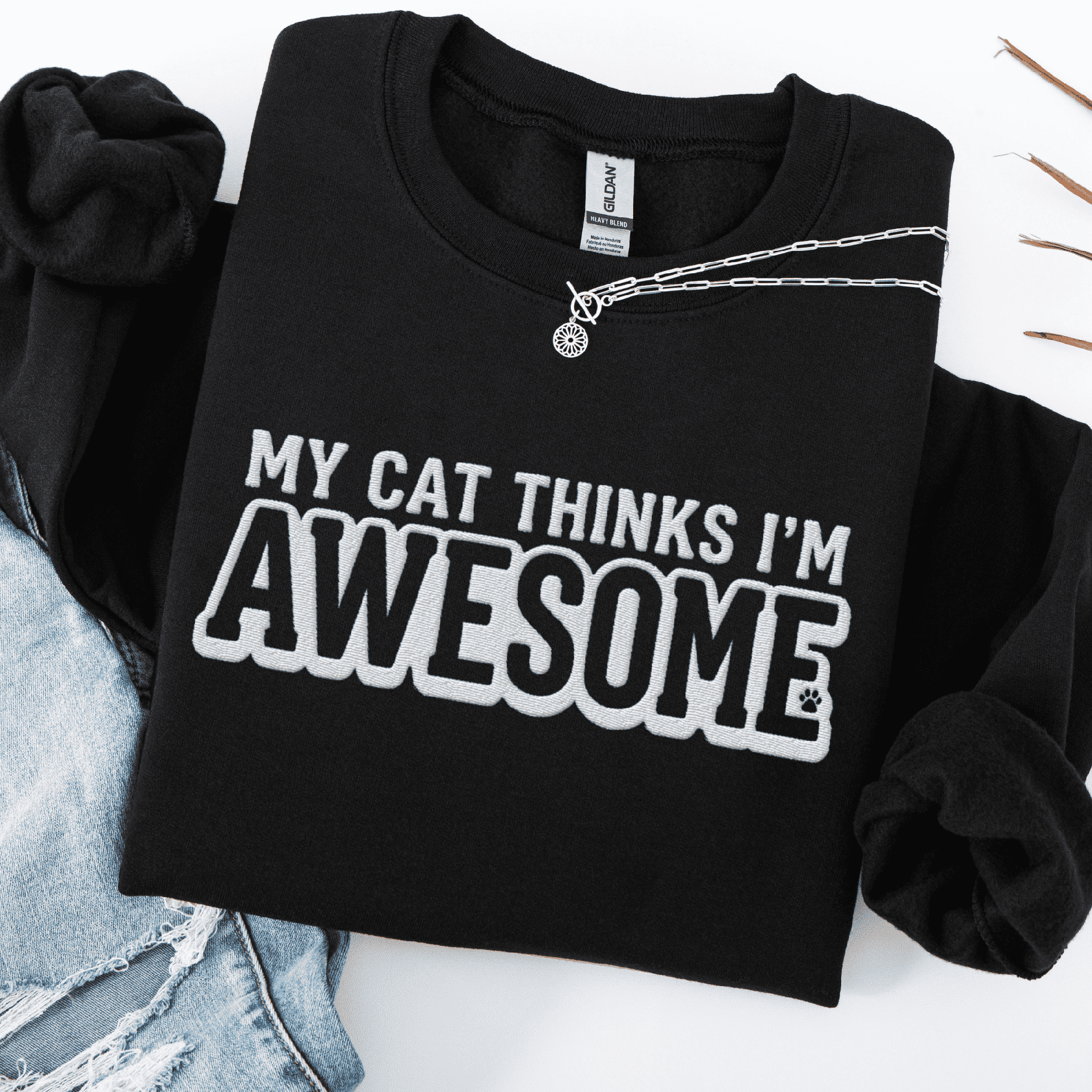 Black cat embroidered sweatshirt with “My Cat Thinks I’m Awesome” text, folded flat on light denim