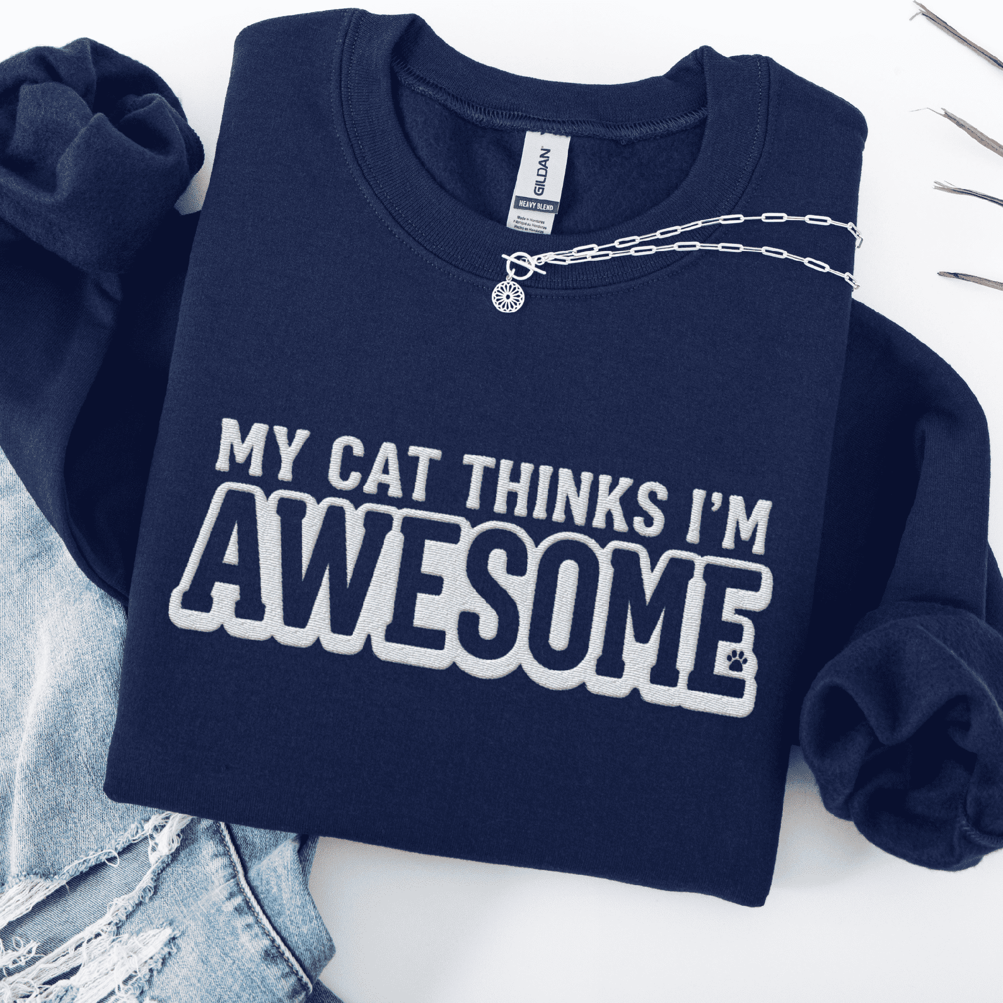 Navy cat embroidered sweatshirt with “My Cat Thinks I’m Awesome” text, folded flat on light denim