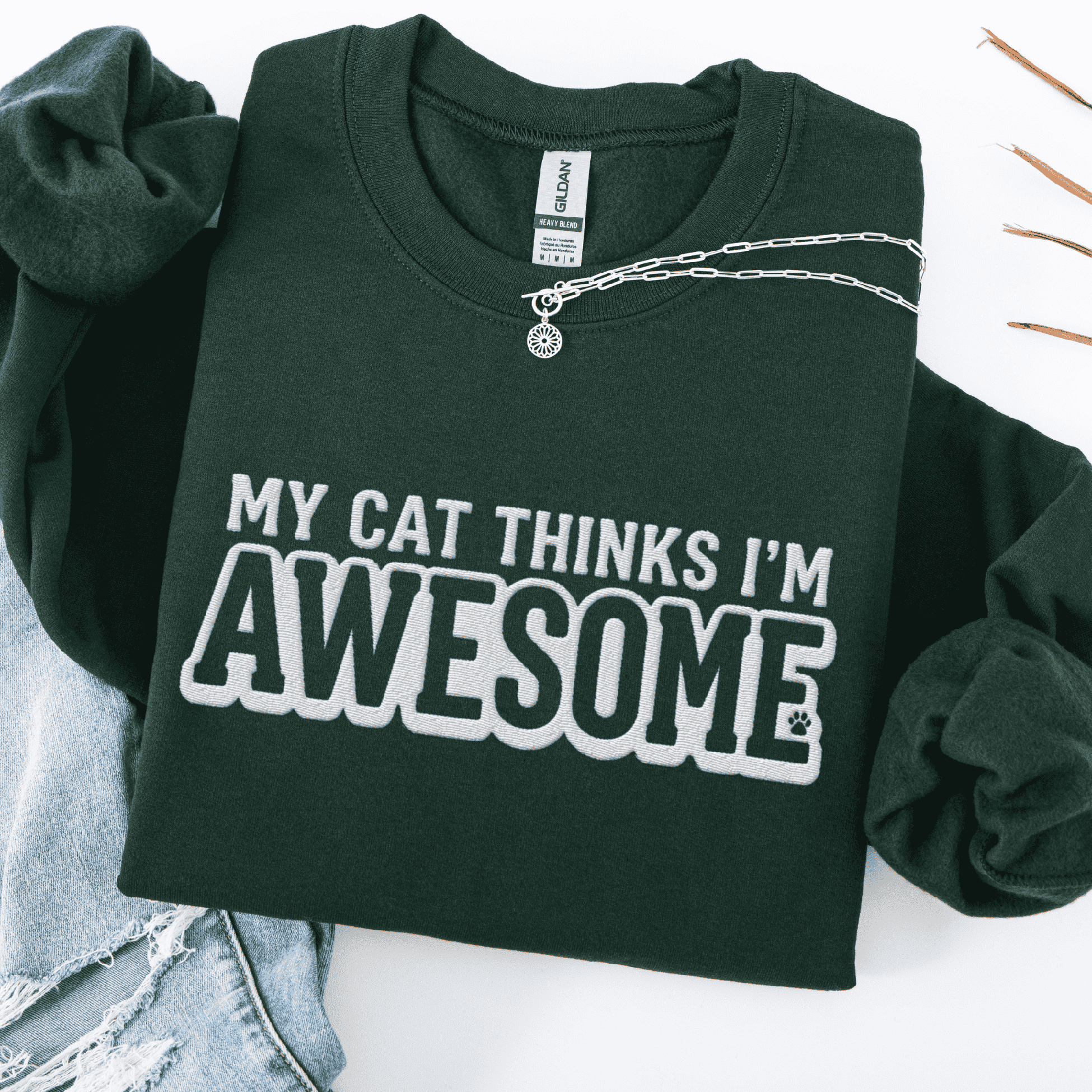 Forest Green cat embroidered sweatshirt with “My Cat Thinks I’m Awesome” text, folded flat on light denim