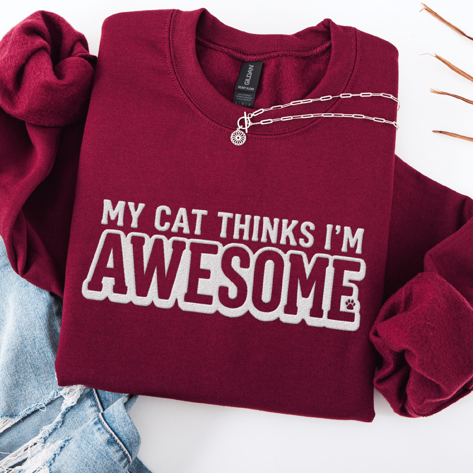 Maroon cat embroidered sweatshirt with “My Cat Thinks I’m Awesome” text, folded flat on light denim