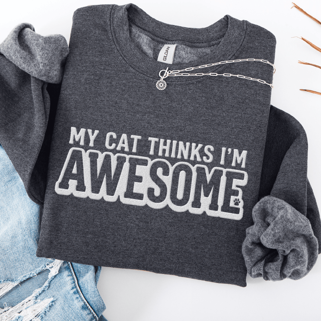 Dark Heather cat embroidered sweatshirt with “My Cat Thinks I’m Awesome” text, folded flat on light denim