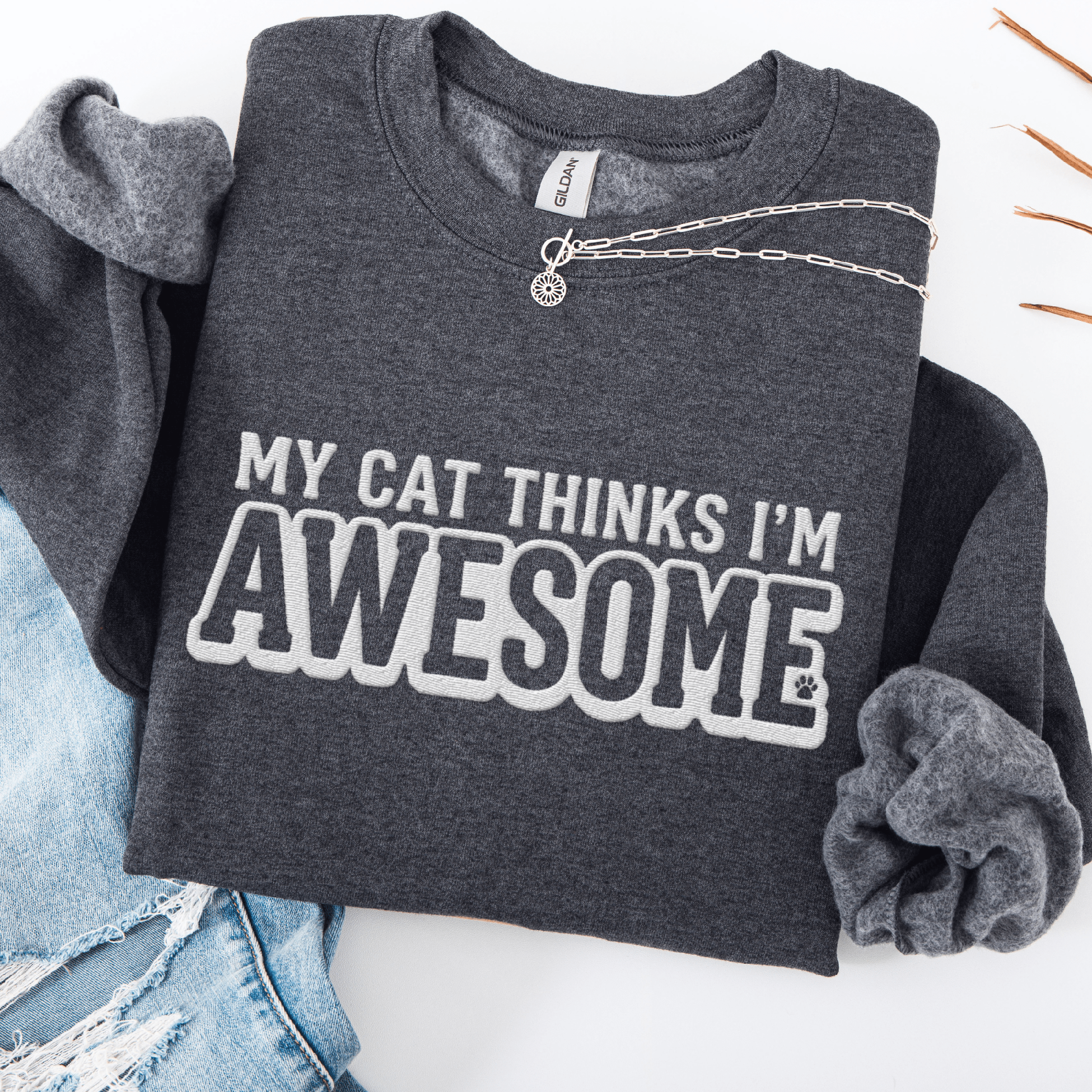 Dark Heather cat embroidered sweatshirt with “My Cat Thinks I’m Awesome” text, folded flat on light denim
