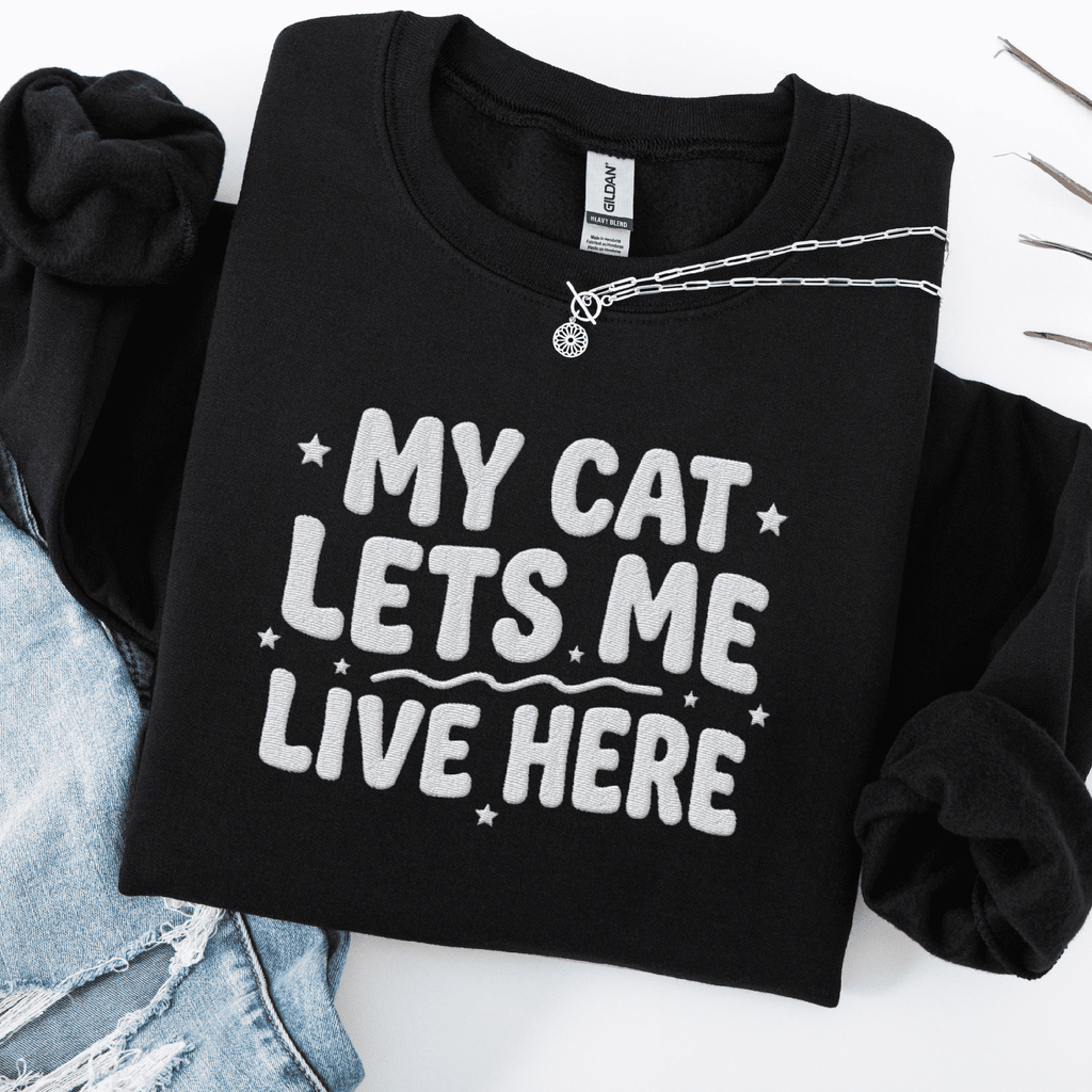 Black cat embroidered sweatshirt with “My Cat Lets Me Live Here” text, folded flat on light denim