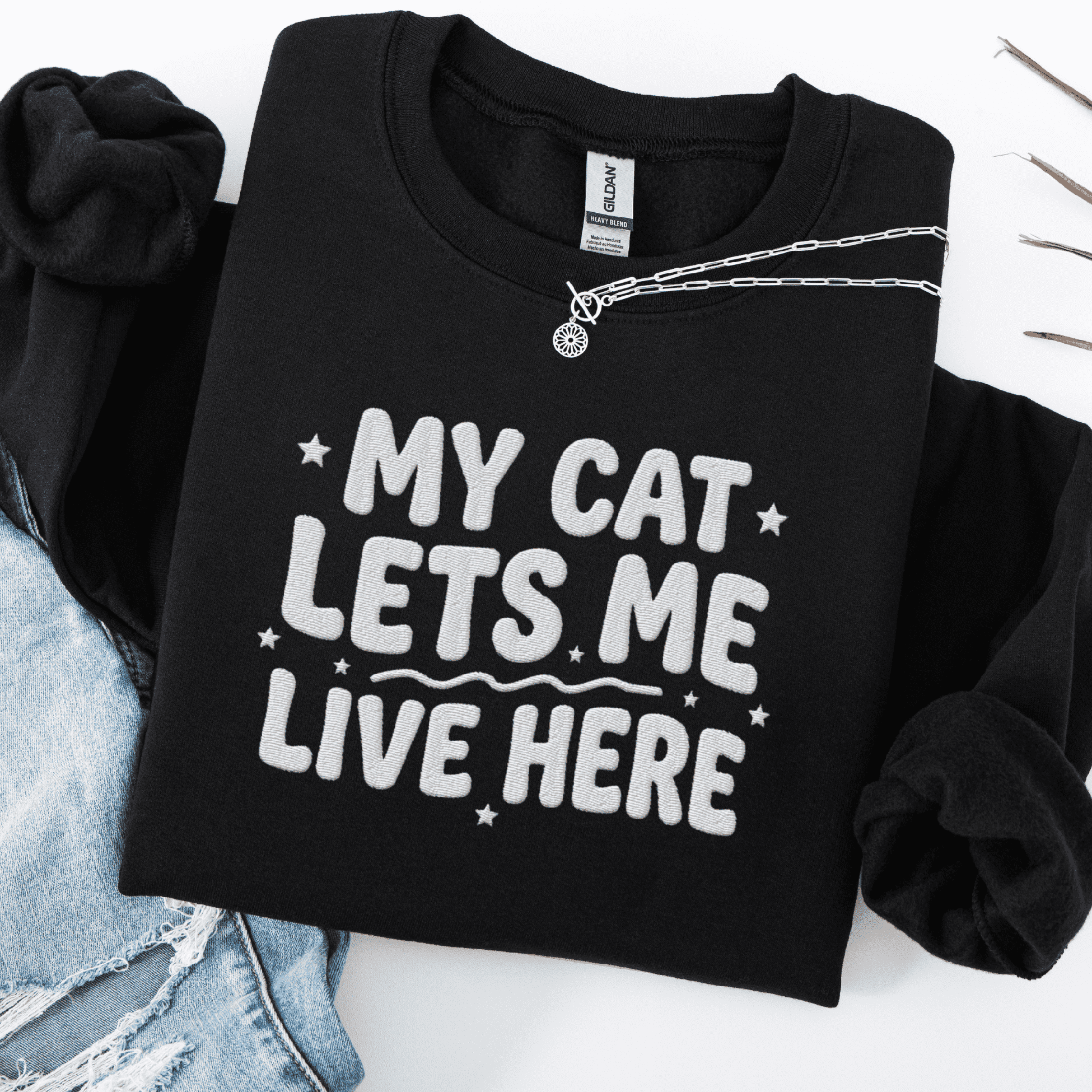 Black cat embroidered sweatshirt with “My Cat Lets Me Live Here” text, folded flat on light denim