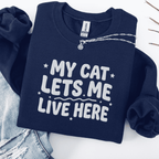 Navy cat embroidered sweatshirt with “My Cat Lets Me Live Here” text, folded flat on light denim