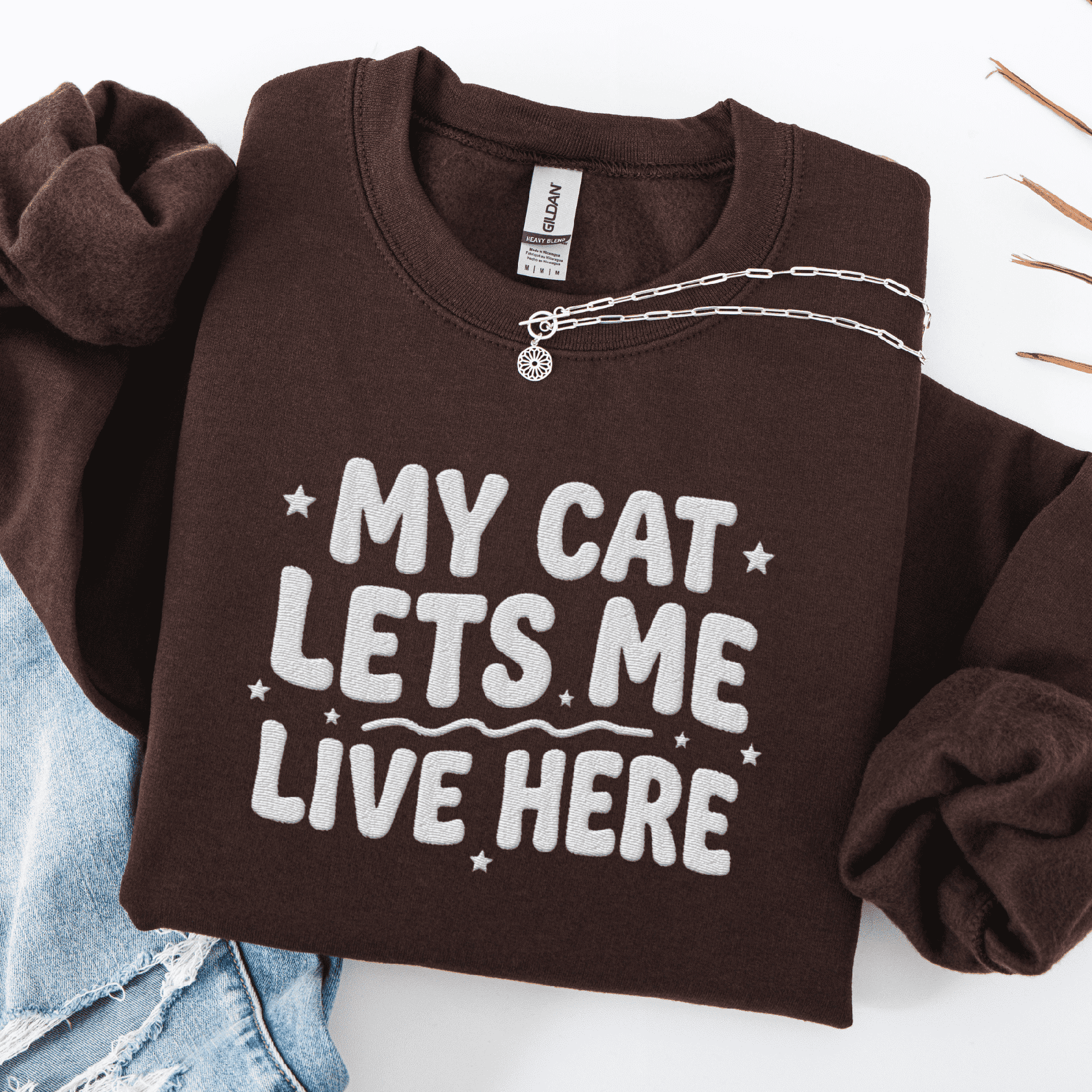 Dark Chocolate cat embroidered sweatshirt with “My Cat Lets Me Live Here” text, folded flat on light denim