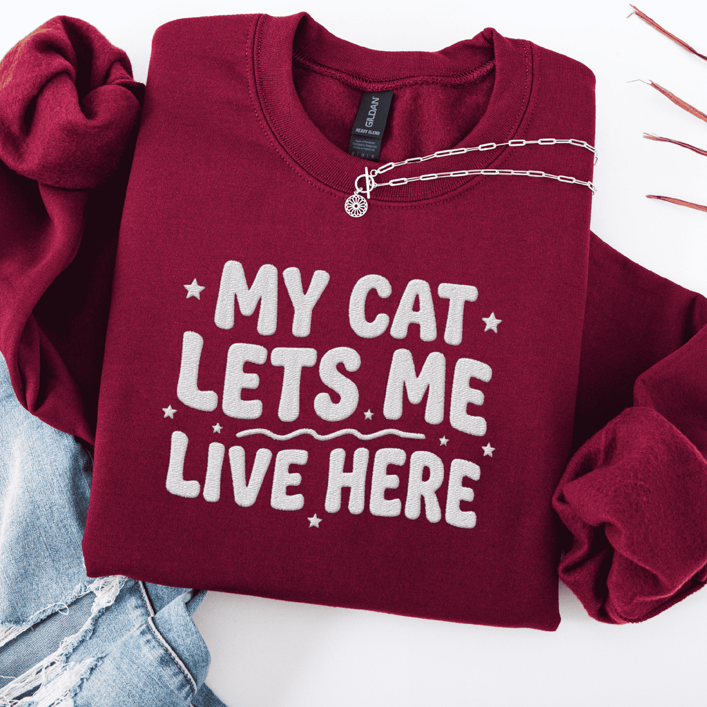 Maroon cat embroidered sweatshirt with “My Cat Lets Me Live Here” text, folded flat on light denim
