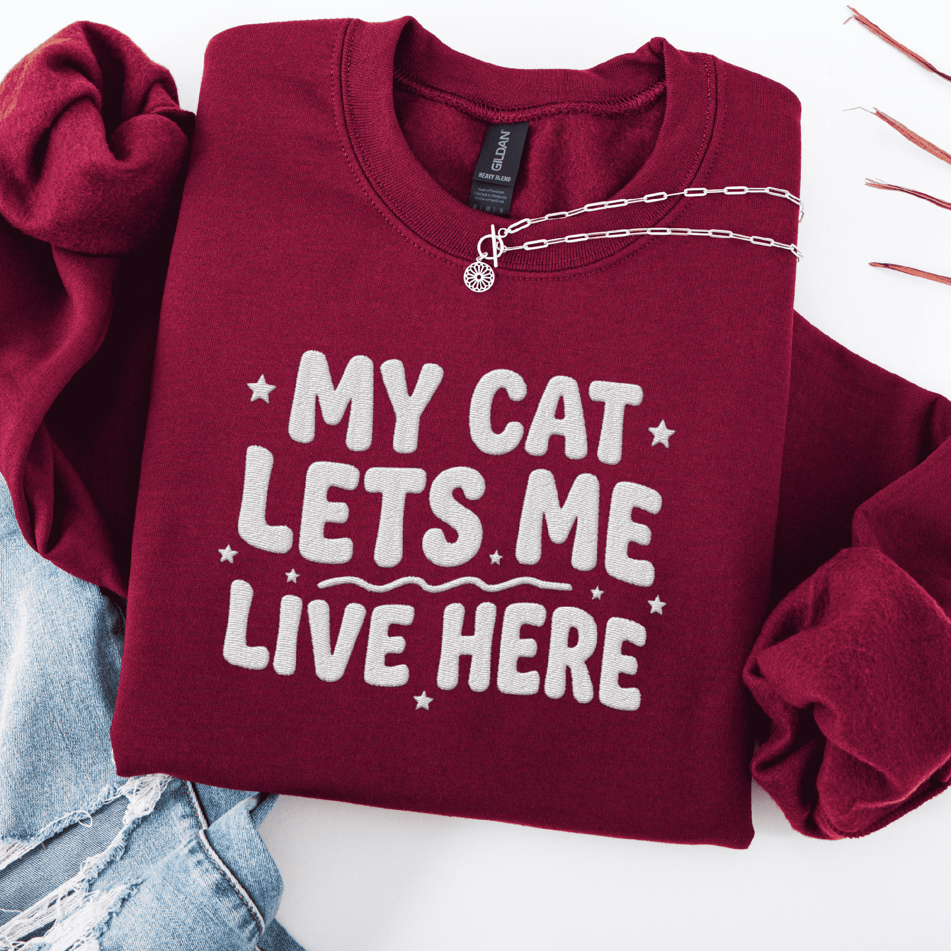 Maroon cat embroidered sweatshirt with “My Cat Lets Me Live Here” text, folded flat on light denim