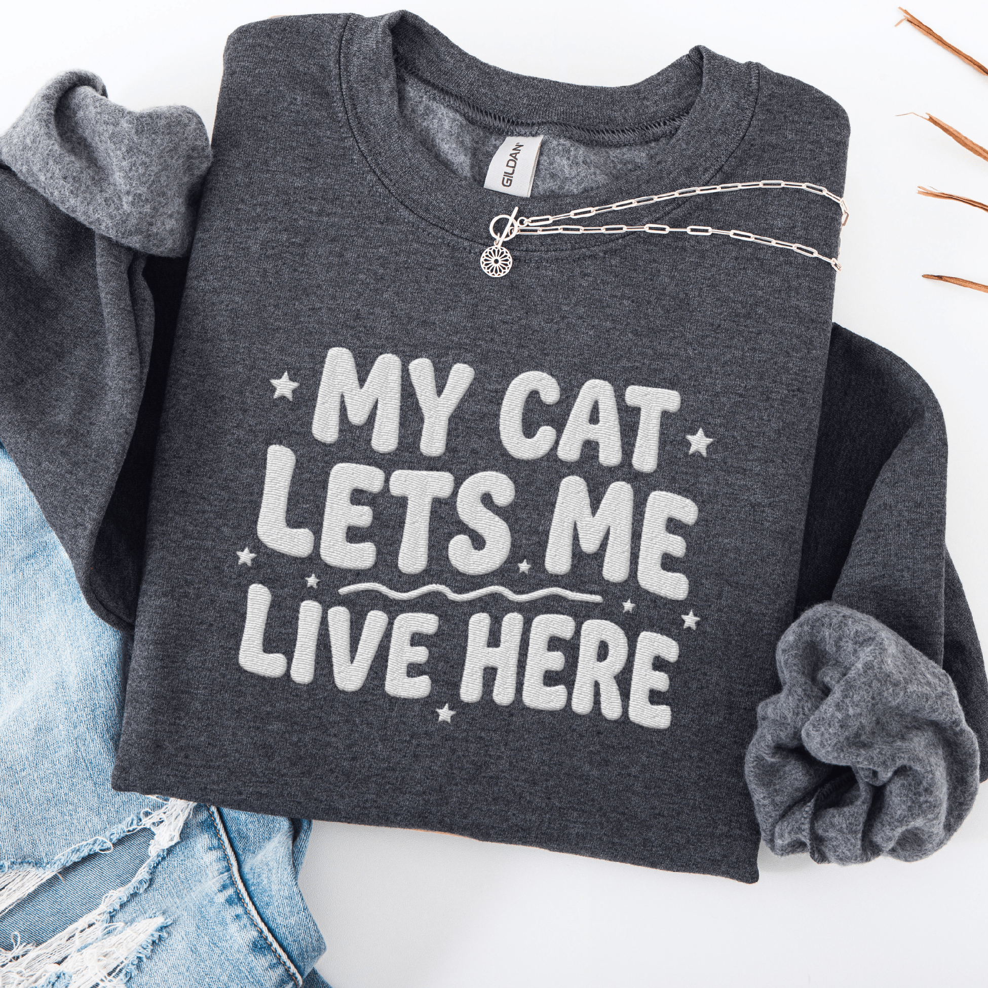 Dark Heather cat embroidered sweatshirt with “My Cat Lets Me Live Here” text, folded flat on light denim