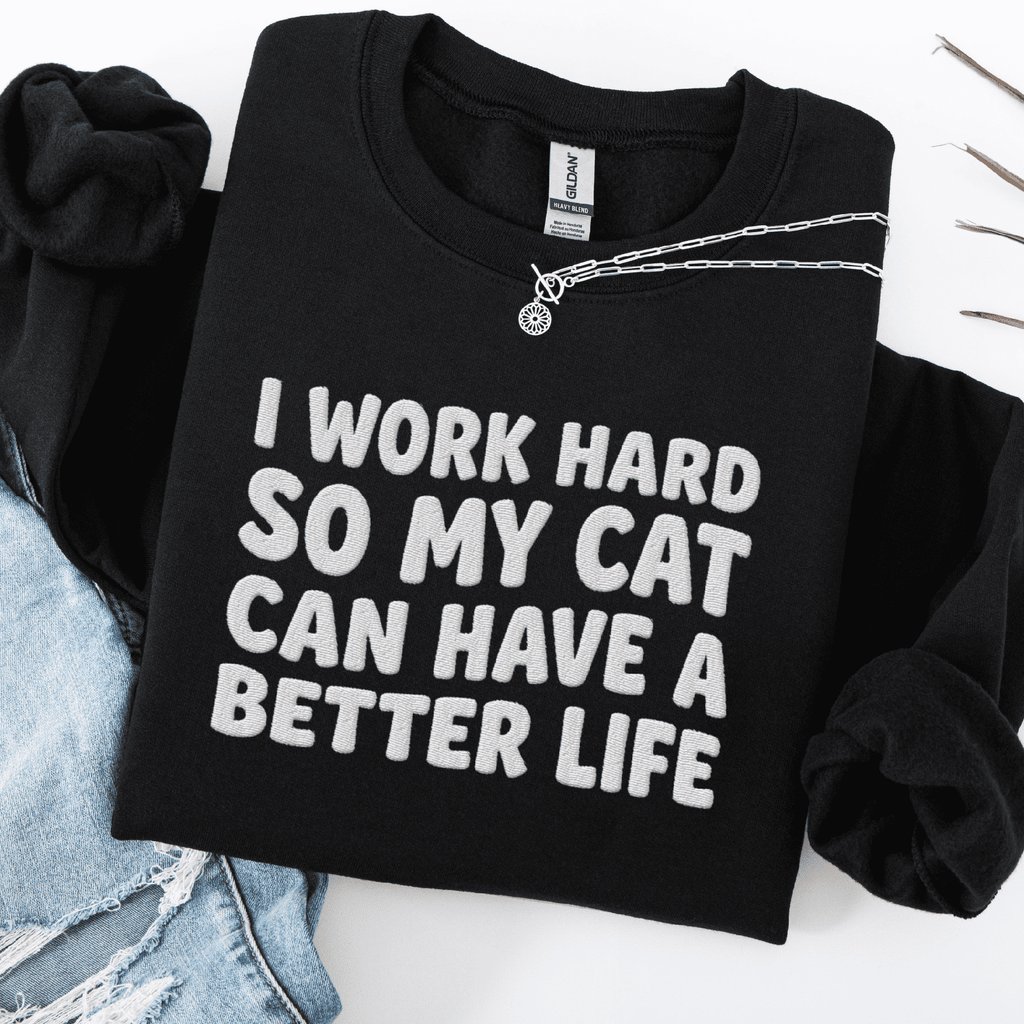 Black cat embroidered sweatshirt with “I Work Hard So My Cat Can Have A Better Life” text, folded flat on light denim