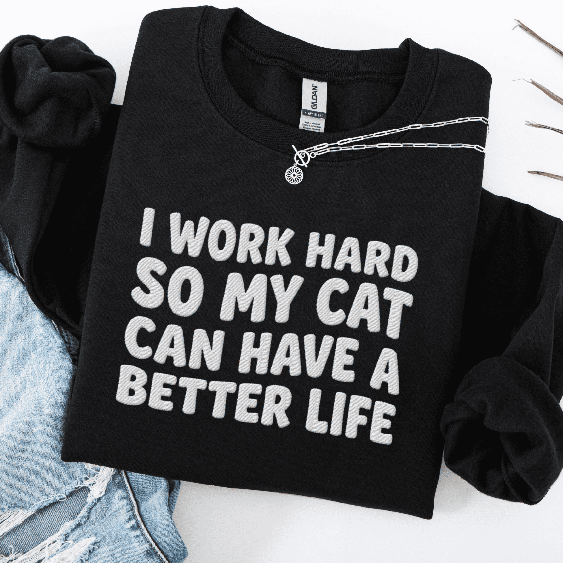 Black cat embroidered sweatshirt with “I Work Hard So My Cat Can Have A Better Life” text, folded flat on light denim