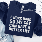 Navy cat embroidered sweatshirt with “I Work Hard So My Cat Can Have A Better Life” text, folded flat on light denim