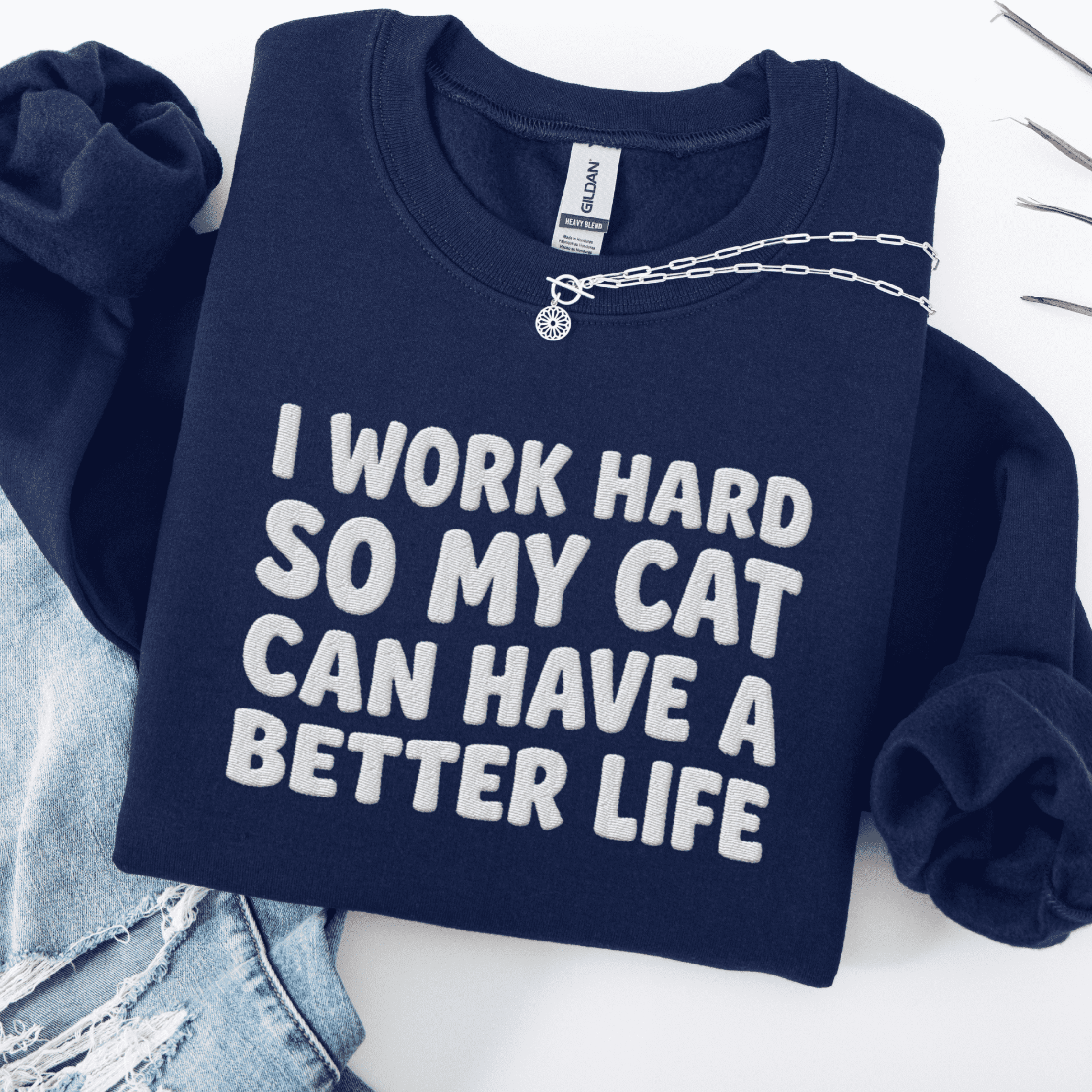Navy cat embroidered sweatshirt with “I Work Hard So My Cat Can Have A Better Life” text, folded flat on light denim