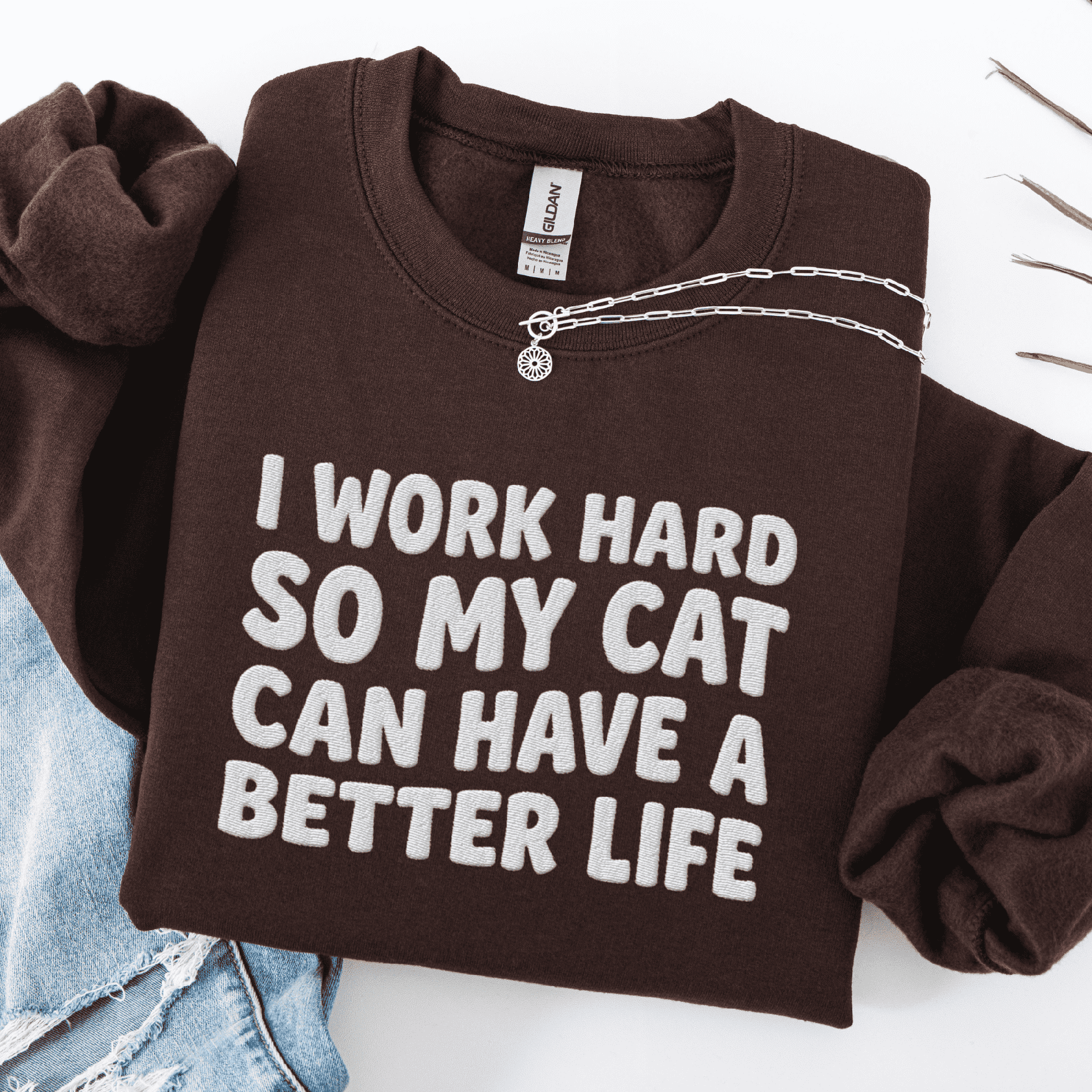Dark Chocolate cat embroidered sweatshirt with “I Work Hard So My Cat Can Have A Better Life” text, folded flat on light denim