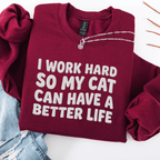 Maroon cat embroidered sweatshirt with “I Work Hard So My Cat Can Have A Better Life” text, folded flat on light denim