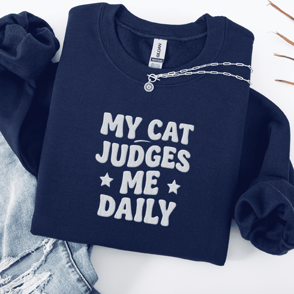 Navy cat embroidered sweatshirt with “My Cat Judges Me Daily” text, folded flat on light denim