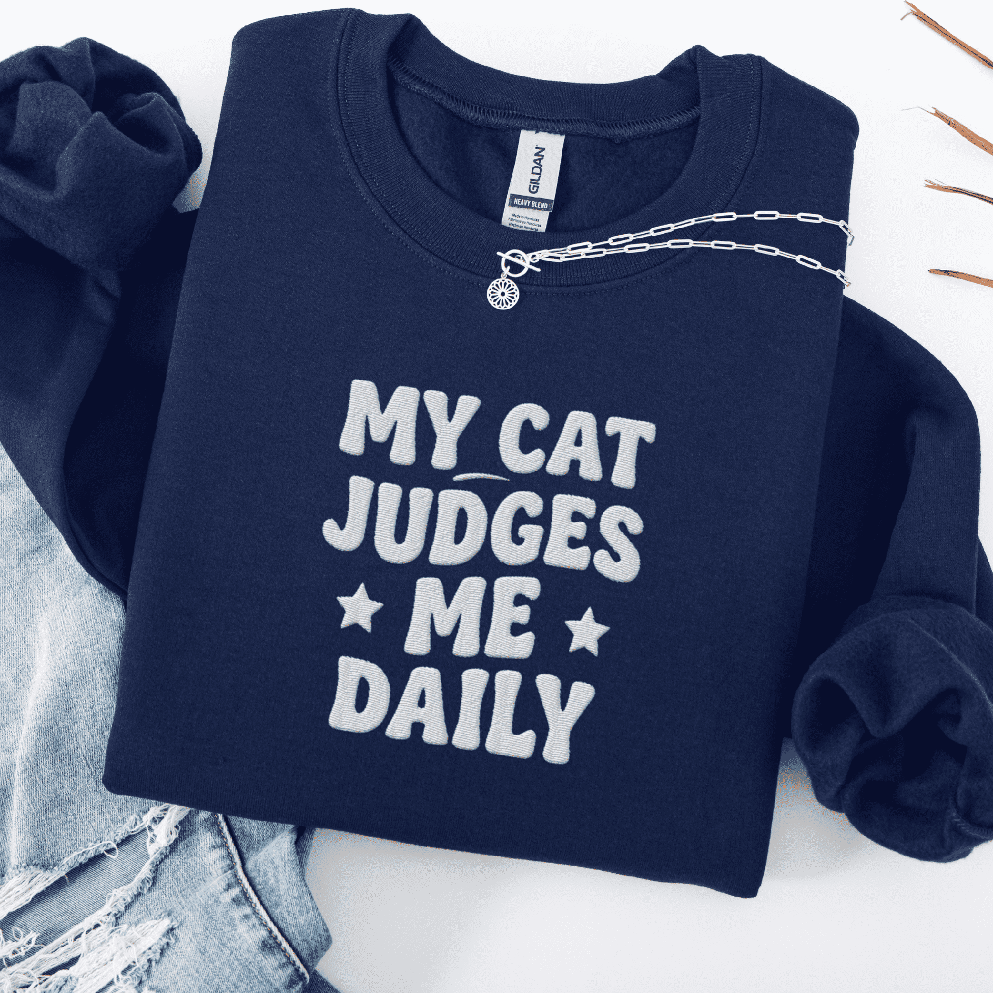 Navy cat embroidered sweatshirt with “My Cat Judges Me Daily” text, folded flat on light denim