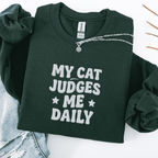 Forest Green cat embroidered sweatshirt with “My Cat Judges Me Daily” text, folded flat on light denim