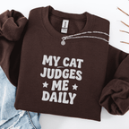 Dark Chocolate cat embroidered sweatshirt with “My Cat Judges Me Daily” text, folded flat on light denim