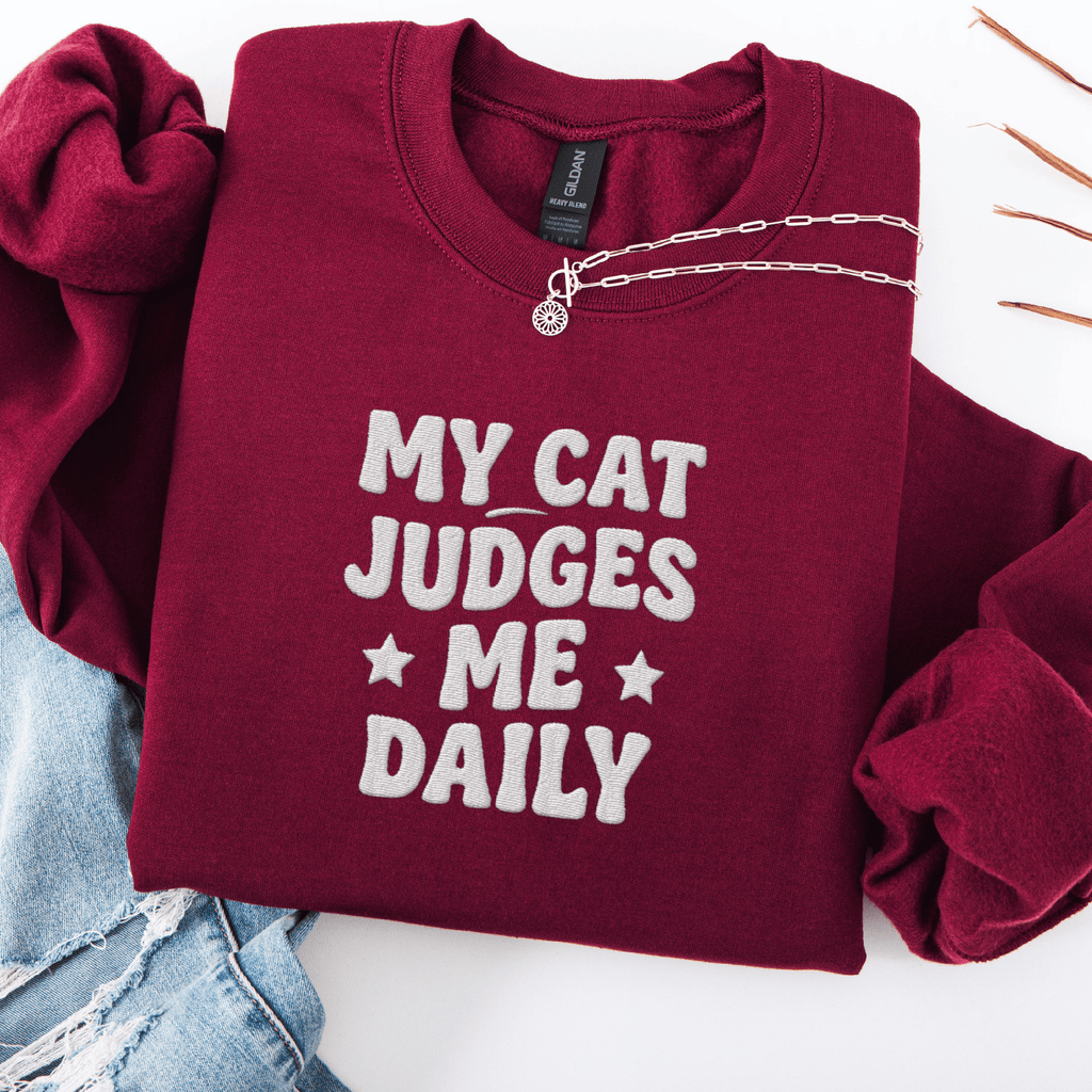 Maroon cat embroidered sweatshirt with “My Cat Judges Me Daily” text, folded flat on light denim