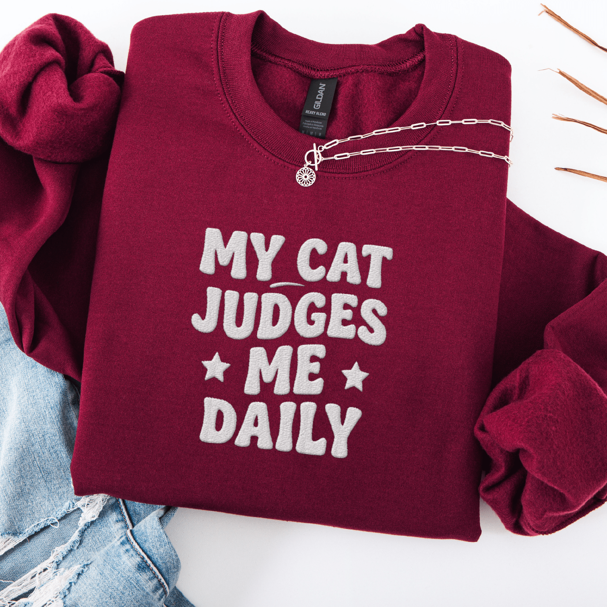 Maroon cat embroidered sweatshirt with “My Cat Judges Me Daily” text, folded flat on light denim