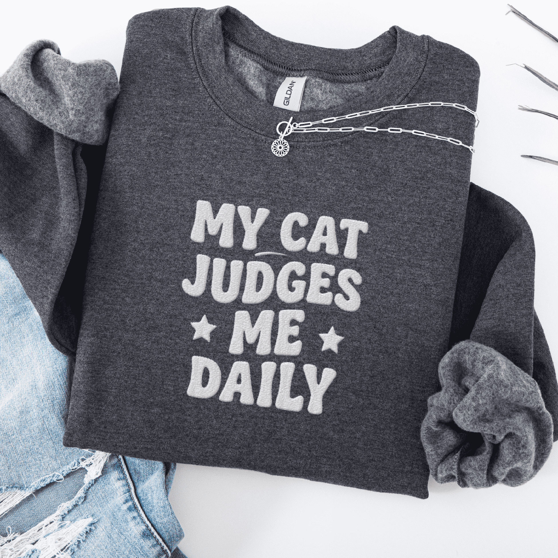 Dark Heather cat embroidered sweatshirt with “My Cat Judges Me Daily” text, folded flat on light denim
