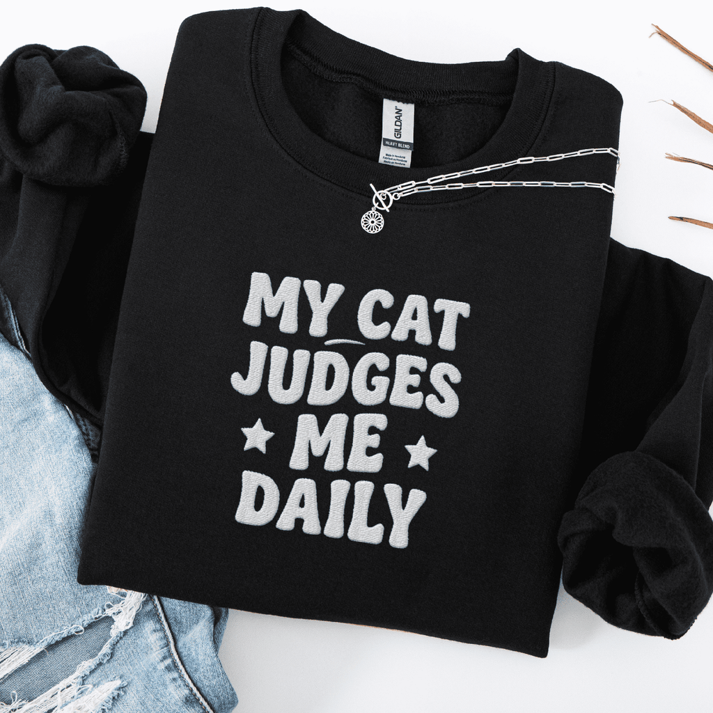 Black cat embroidered sweatshirt with “My Cat Judges Me Daily” text, folded flat on light denim