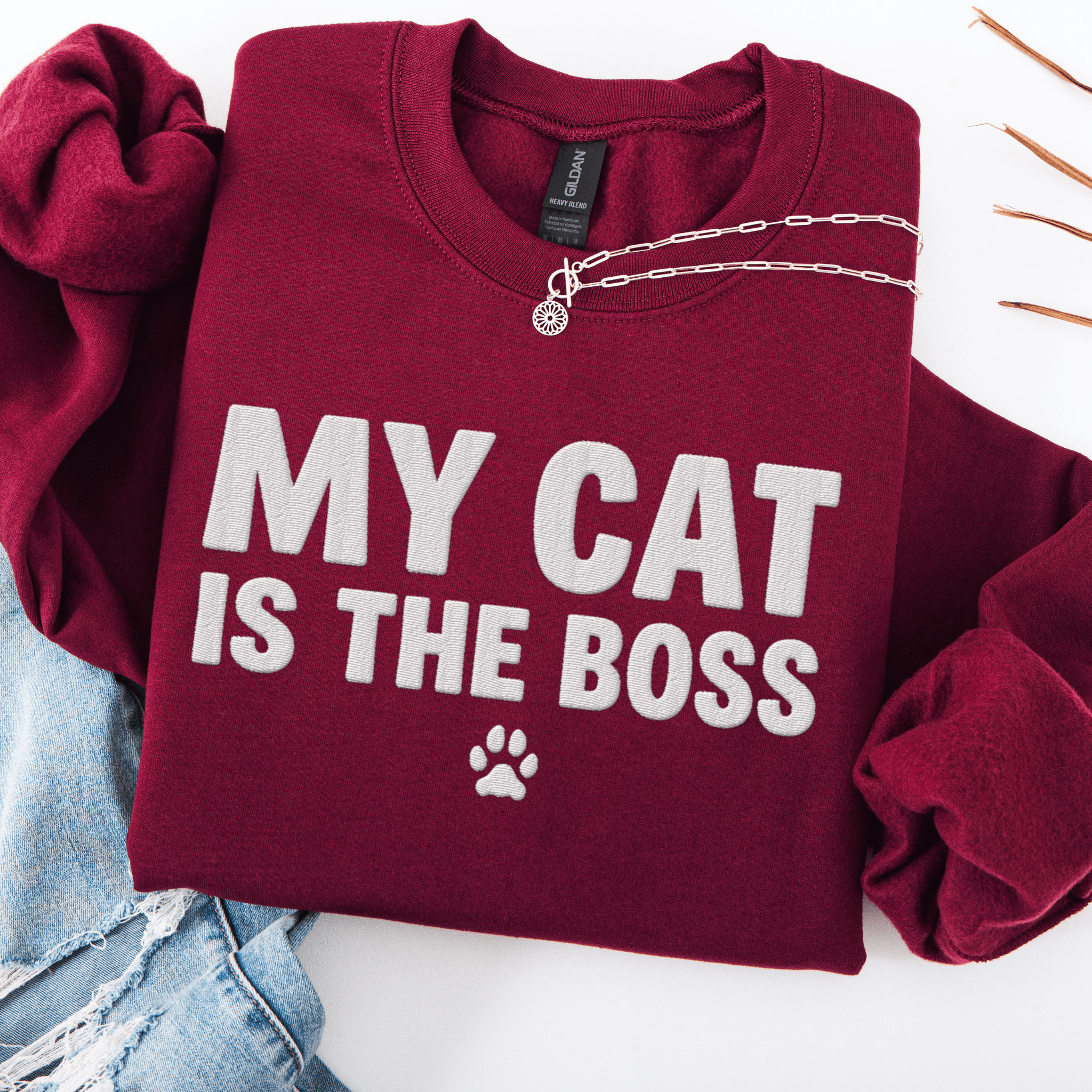 Maroon cat embroidered sweatshirt with “My Cat Is The Boss” text, folded flat on light denim