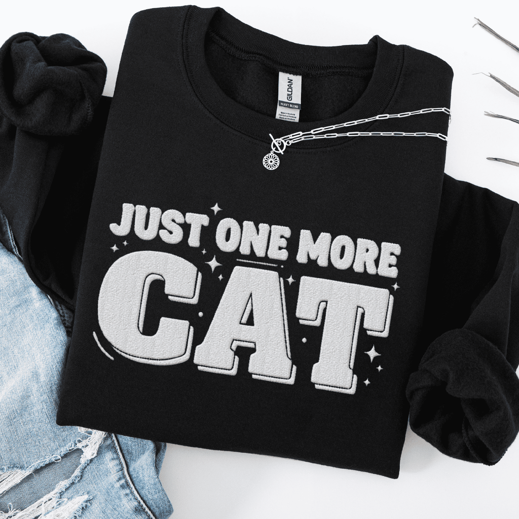 Black one more cat embroidered sweatshirt with “Just One More Cat” text, folded flat on light denim