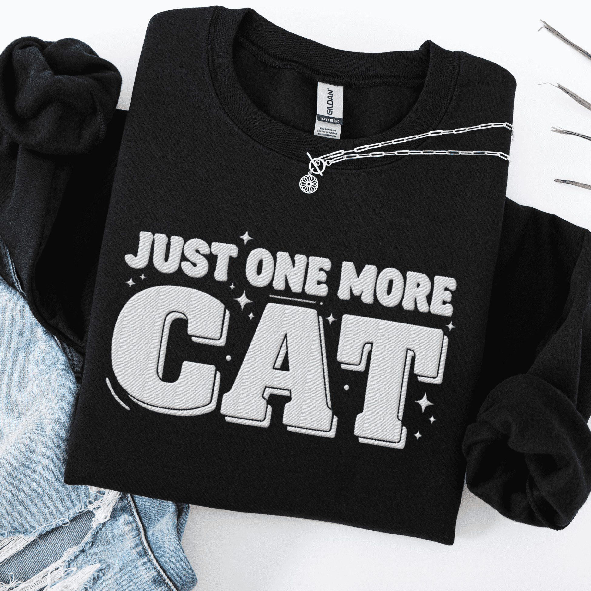 Black one more cat embroidered sweatshirt with “Just One More Cat” text, folded flat on light denim