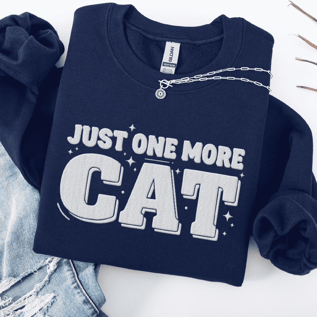 Navy one more cat embroidered sweatshirt with “Just One More Cat” text, folded flat on light denim