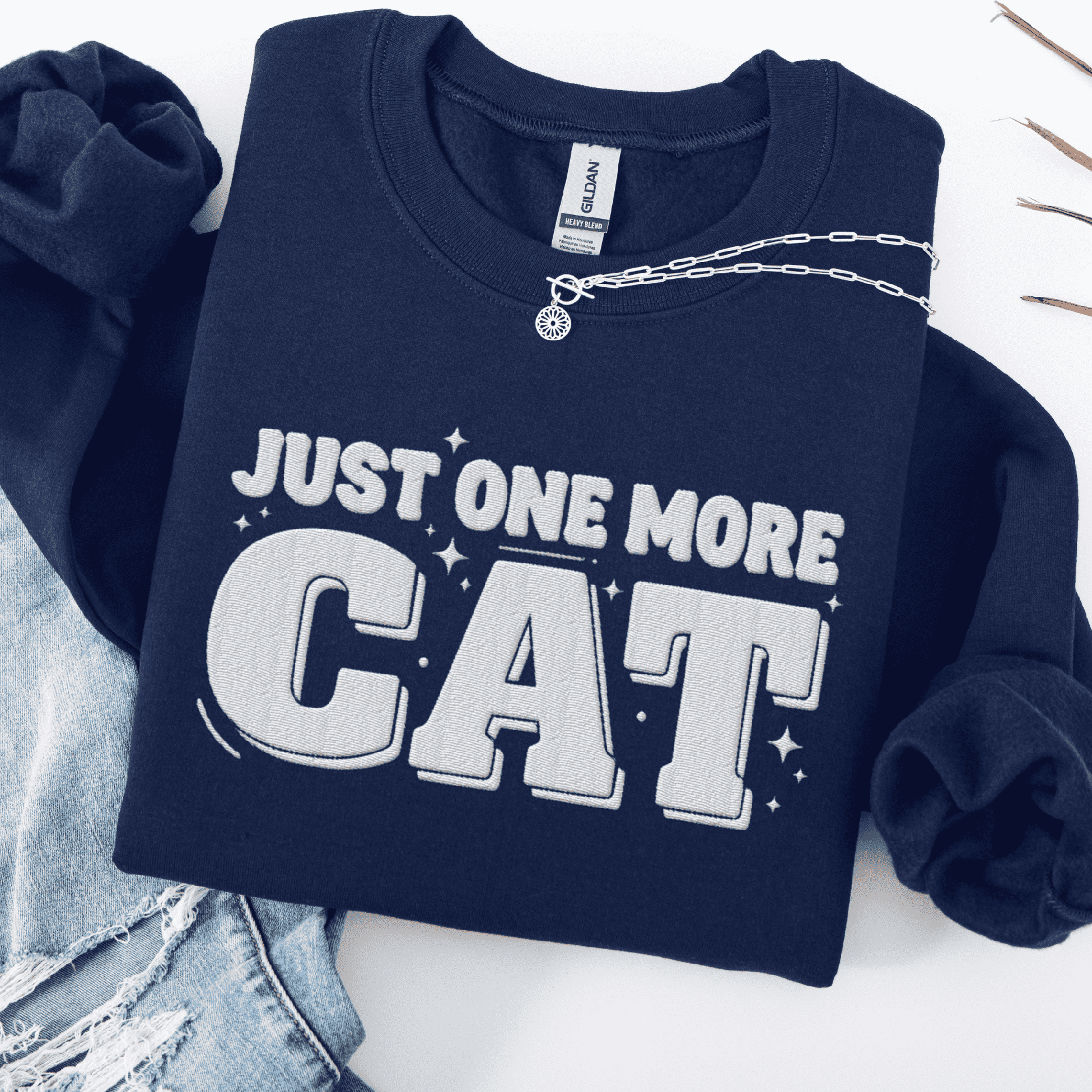 Navy one more cat embroidered sweatshirt with “Just One More Cat” text, folded flat on light denim