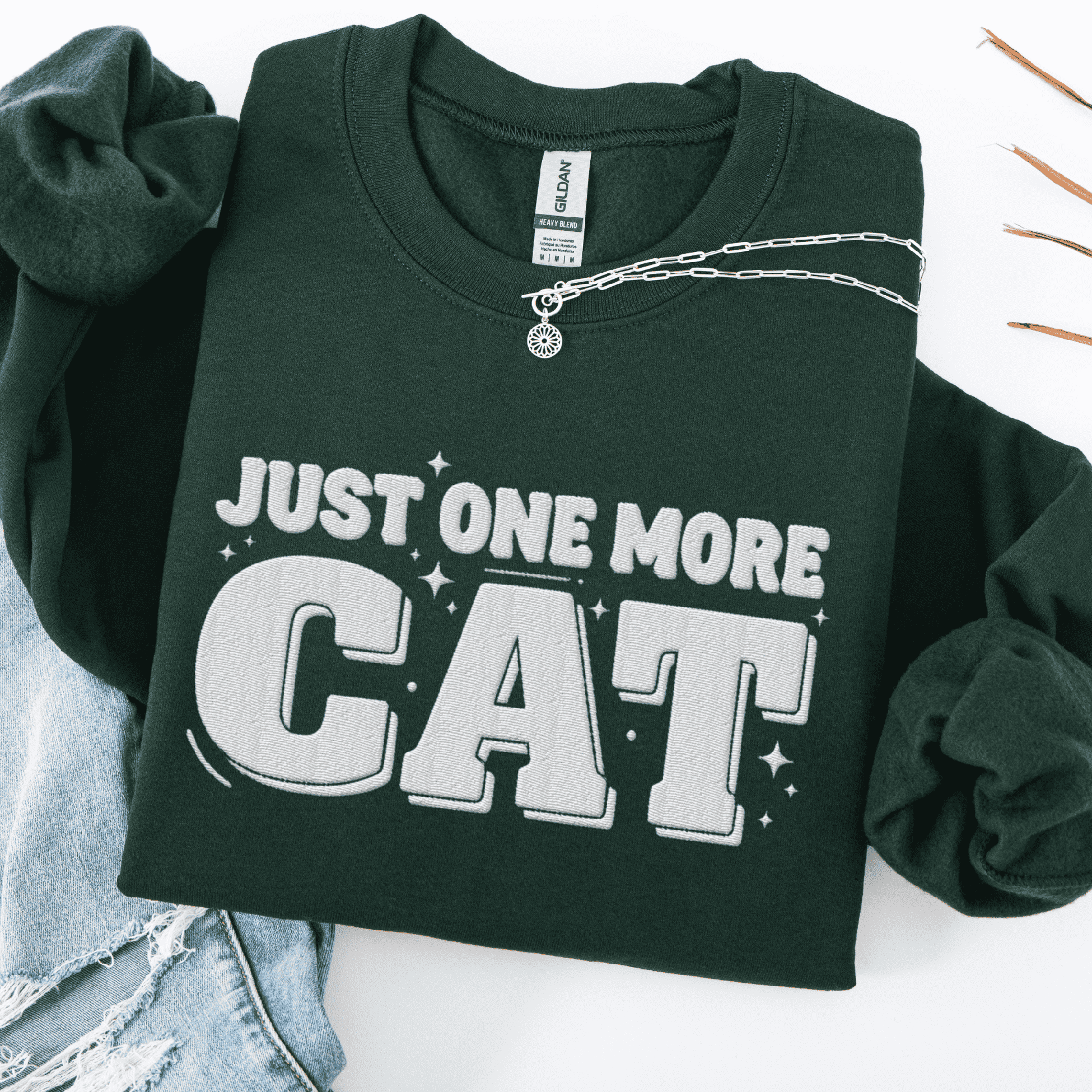 Forest Green one more cat embroidered sweatshirt with “Just One More Cat” text, folded flat on light denim