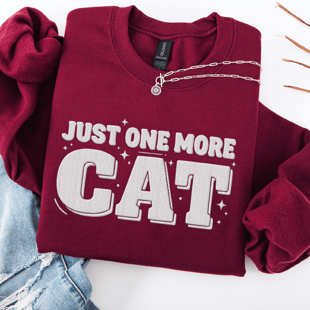 Maroon one more cat embroidered sweatshirt with “Just One More Cat” text, folded flat on light denim