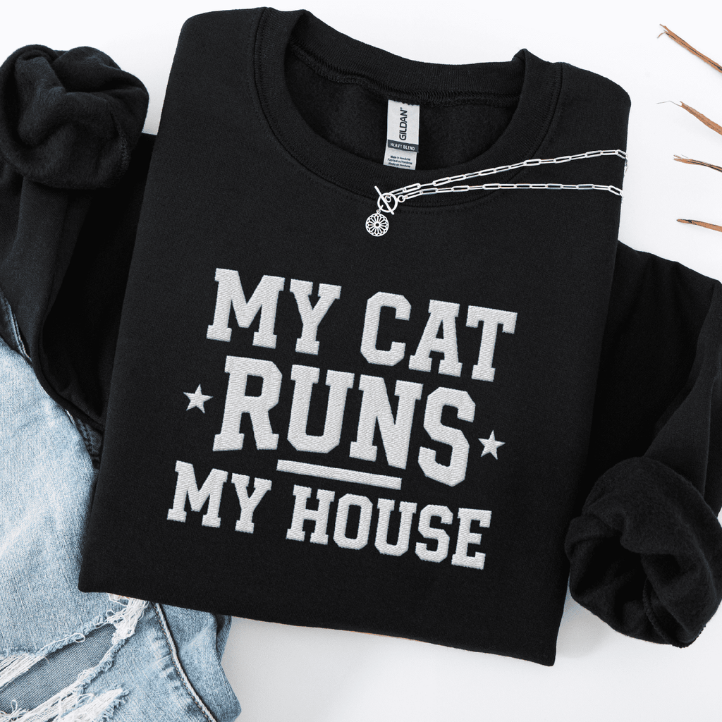 Black cat embroidered sweatshirt with “My Cat Runs My House” text, folded flat on light denim