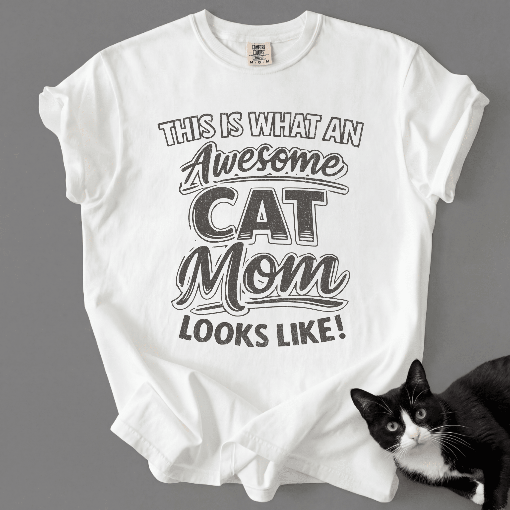 White cat mom t-shirt with “This Is What an Awesome Cat Mom Looks Like” graphic shown with a black cat at the bottom corner