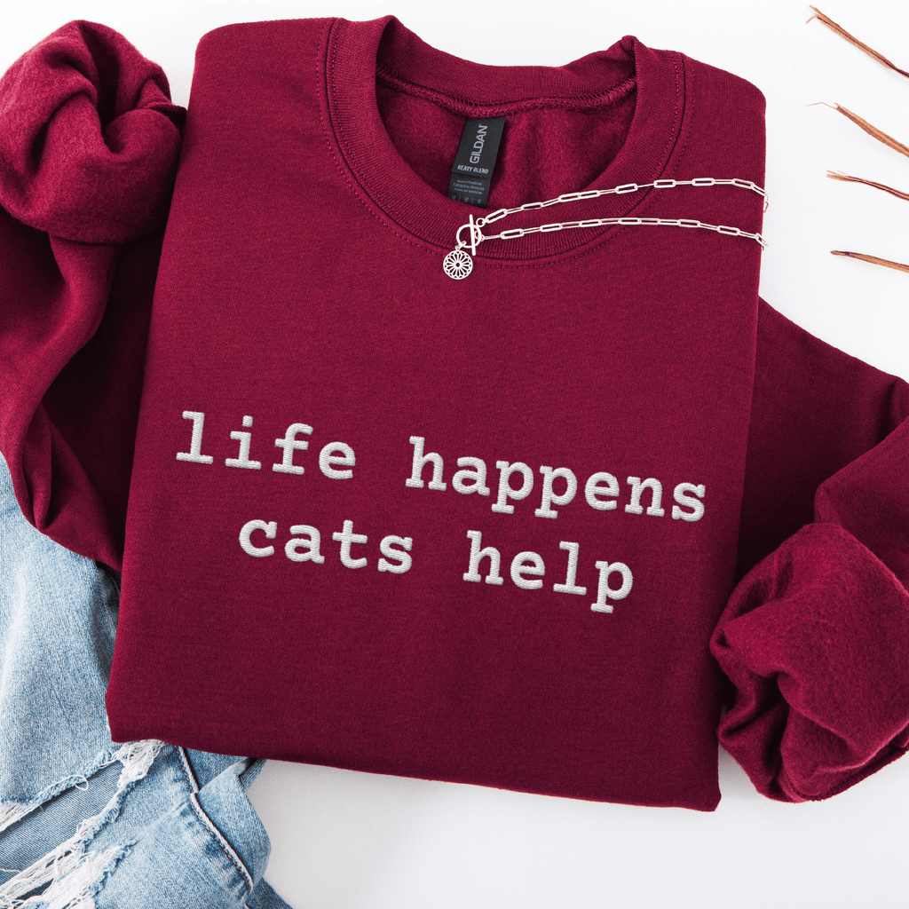 Maroon embroidered cat sweatshirt with the phrase “life happens cats help” shown folded flat