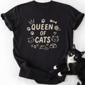 Black Queen of Cats cat t shirt with cat-themed doodle icons shown laid flat with a tuxedo cat beside it