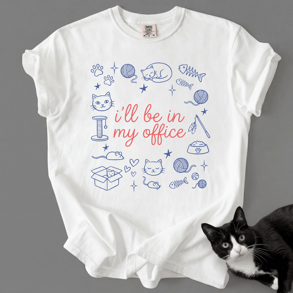 White funny cat t shirt with I'll be in my office text and pastel cat office illustration, shown flat with a black cat beside it
