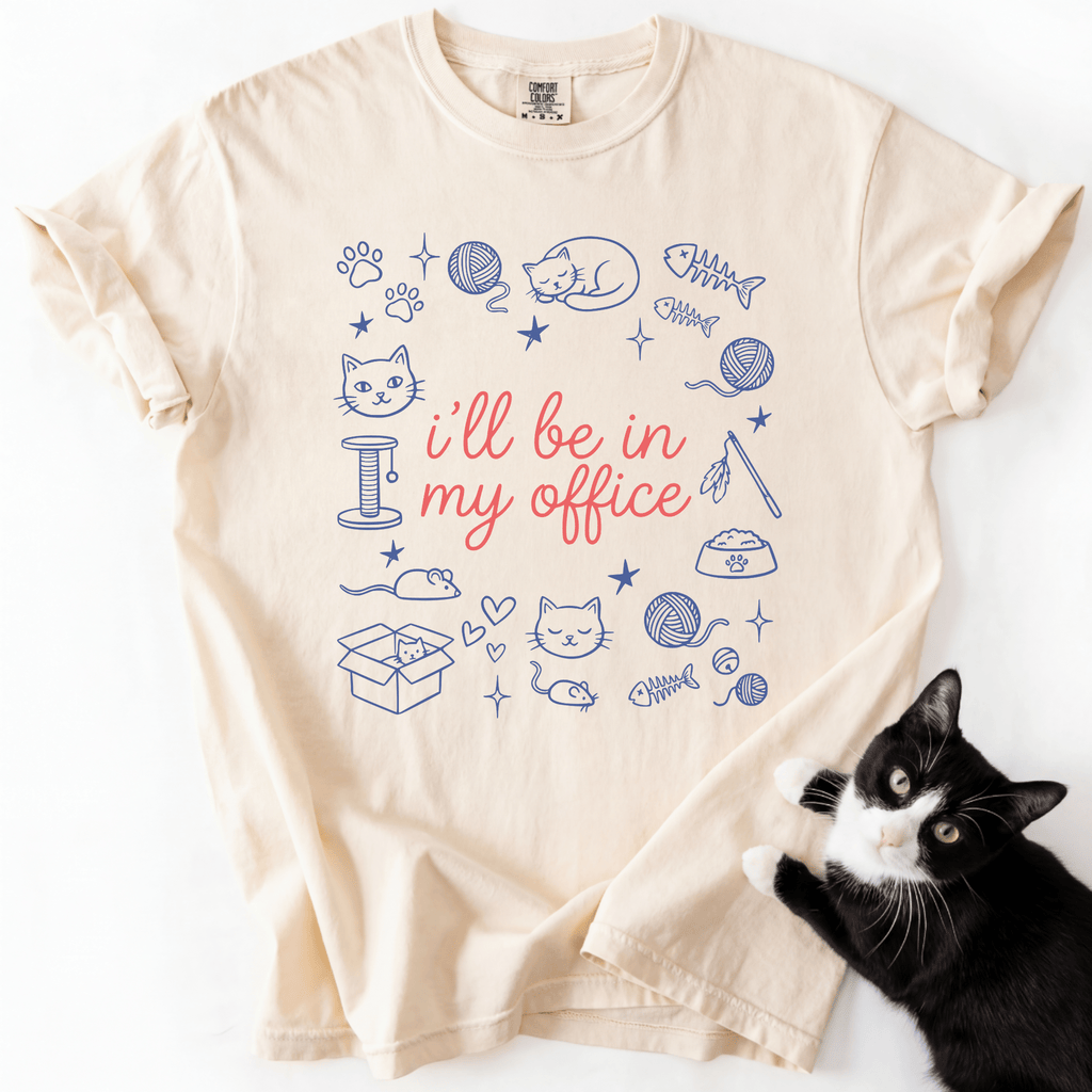 Ivory funny cat t shirt with I'll be in my office text and pastel cat themed office doodles, shown flat with a black cat beside it