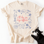 Ivory funny cat t shirt with I'll be in my office text and pastel cat themed office doodles, shown flat with a black cat beside it