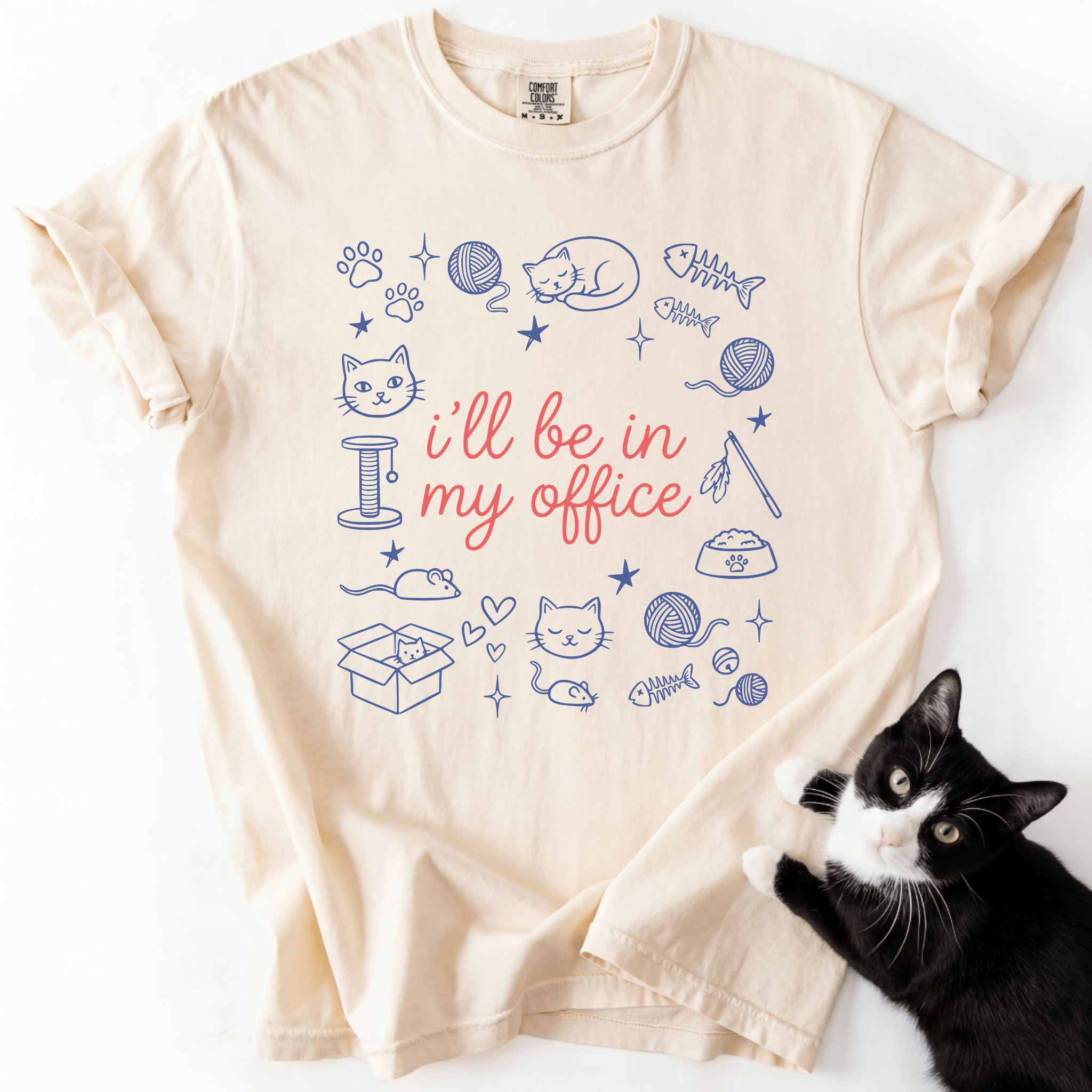 Ivory funny cat t shirt with I'll be in my office text and pastel cat themed office doodles, shown flat with a black cat beside it