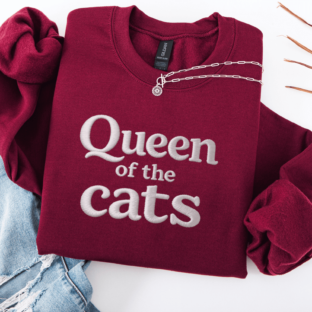 Maroon cat embroidered sweatshirt with Queen of the Cats text folded flat with jeans