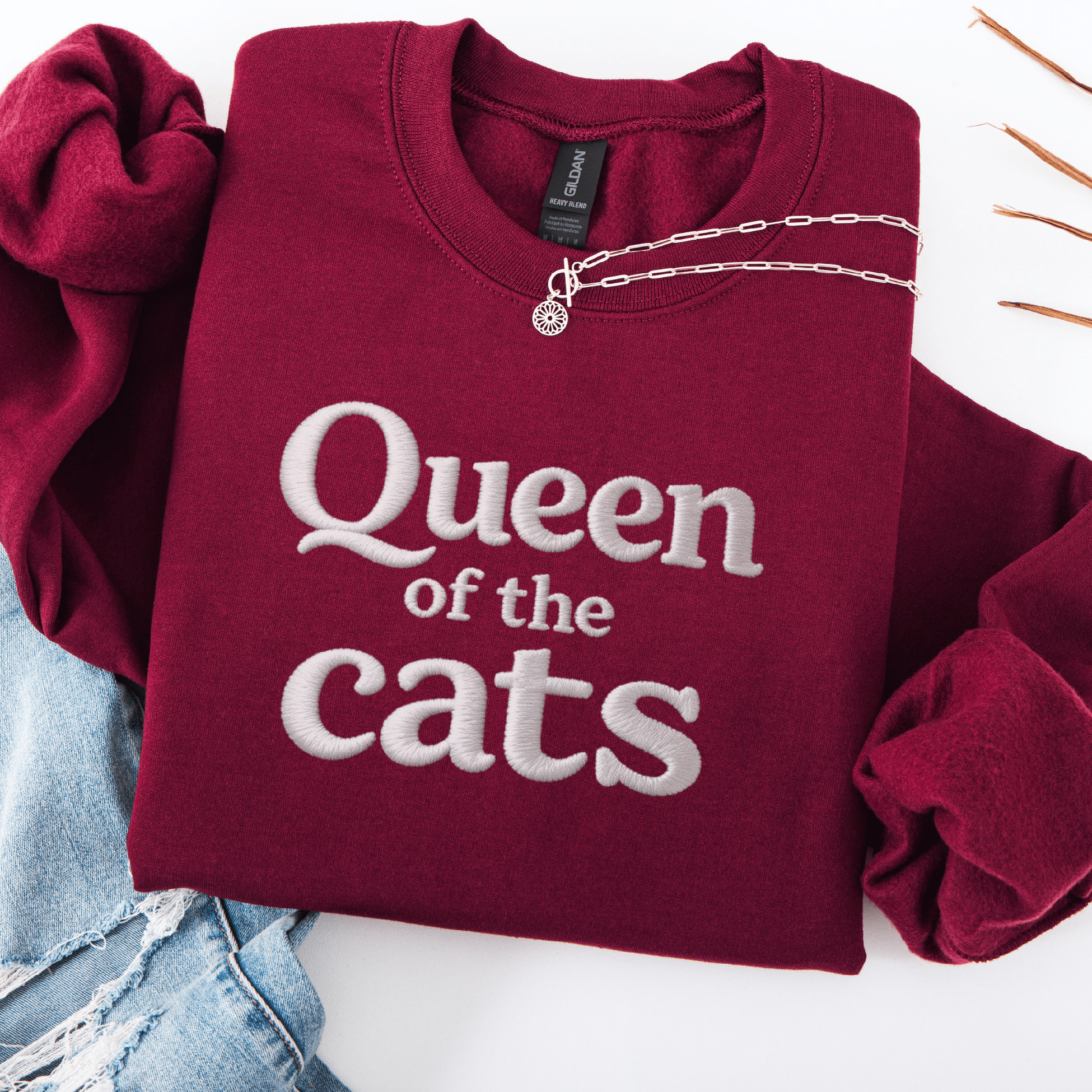 Maroon cat embroidered sweatshirt with Queen of the Cats text folded flat with jeans